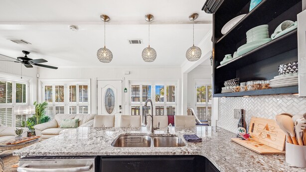 A kitchen with a granite countertop, stainless steel sink, and black cabinets. There are hanging pendant lights, a ceiling fan, and a built-in shelf with dishes. - Steps to Ocean! PCB Family Home w/ Outdoor Oasis (Panama City Beach)