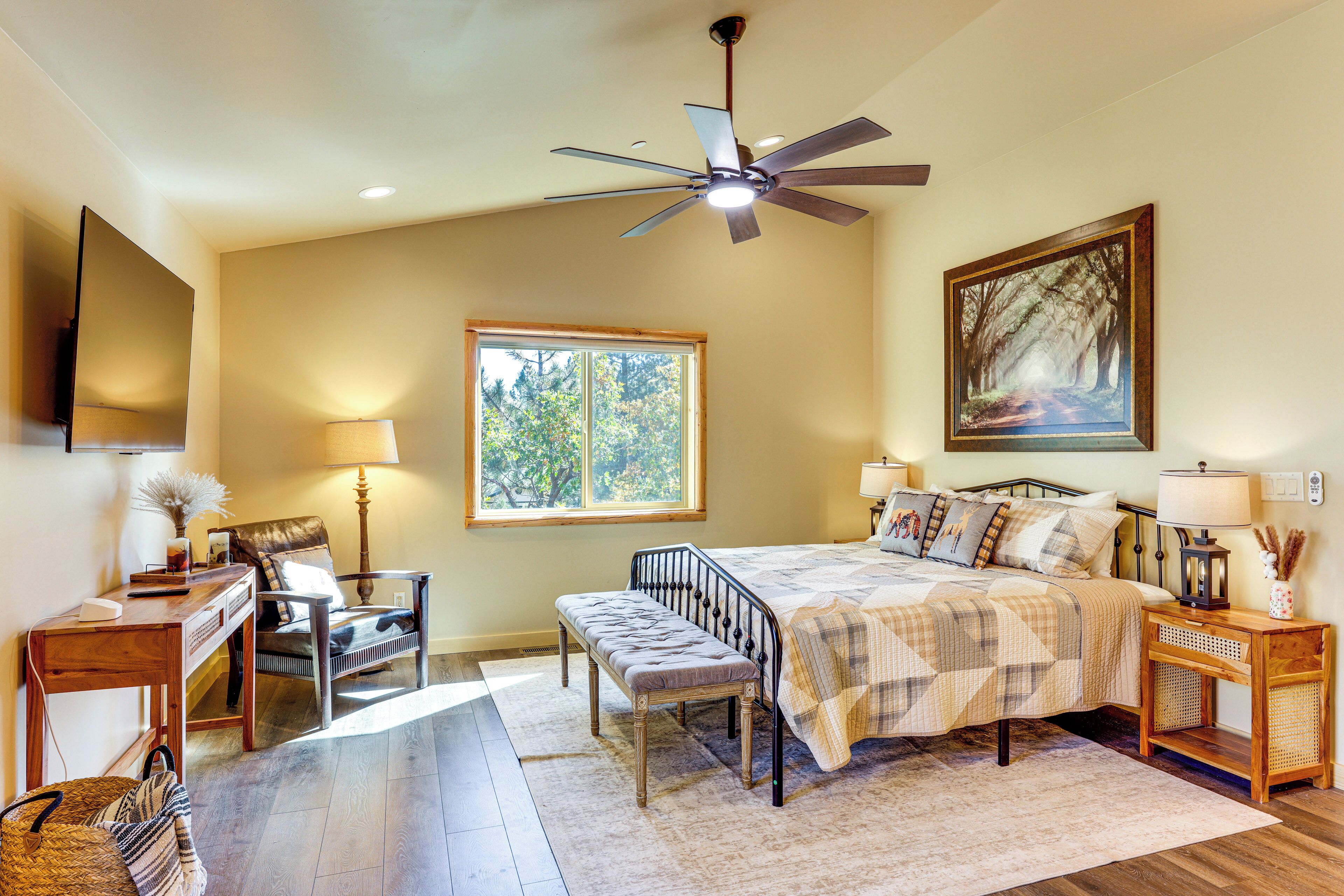 A bedroom with a bed, a desk, a chair, a bench, a window, a ceiling fan, and a painting.