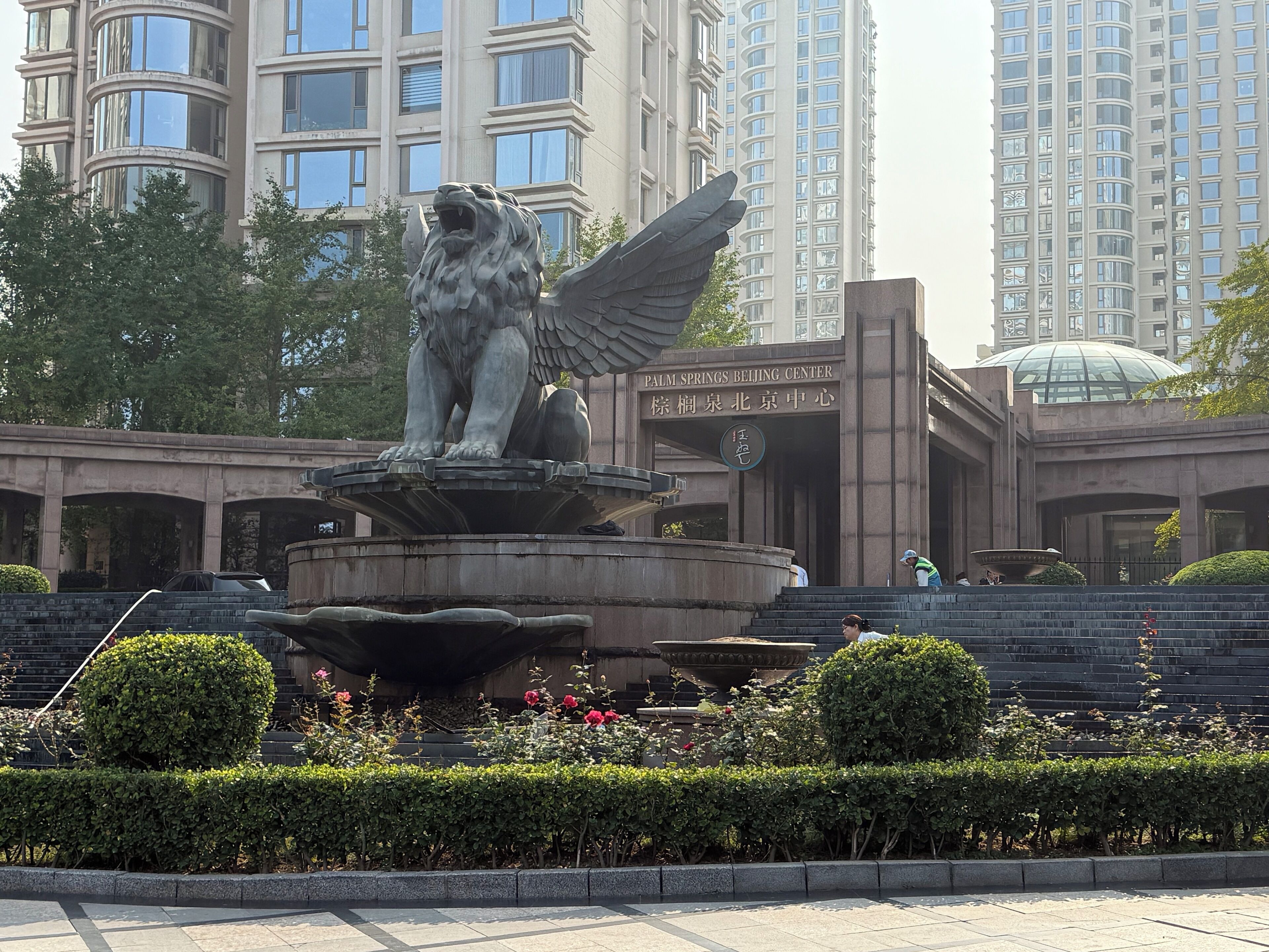 A stone lion statue with wings, a fountain, and a building with a sign reading "Palm Springs Beijing Center".