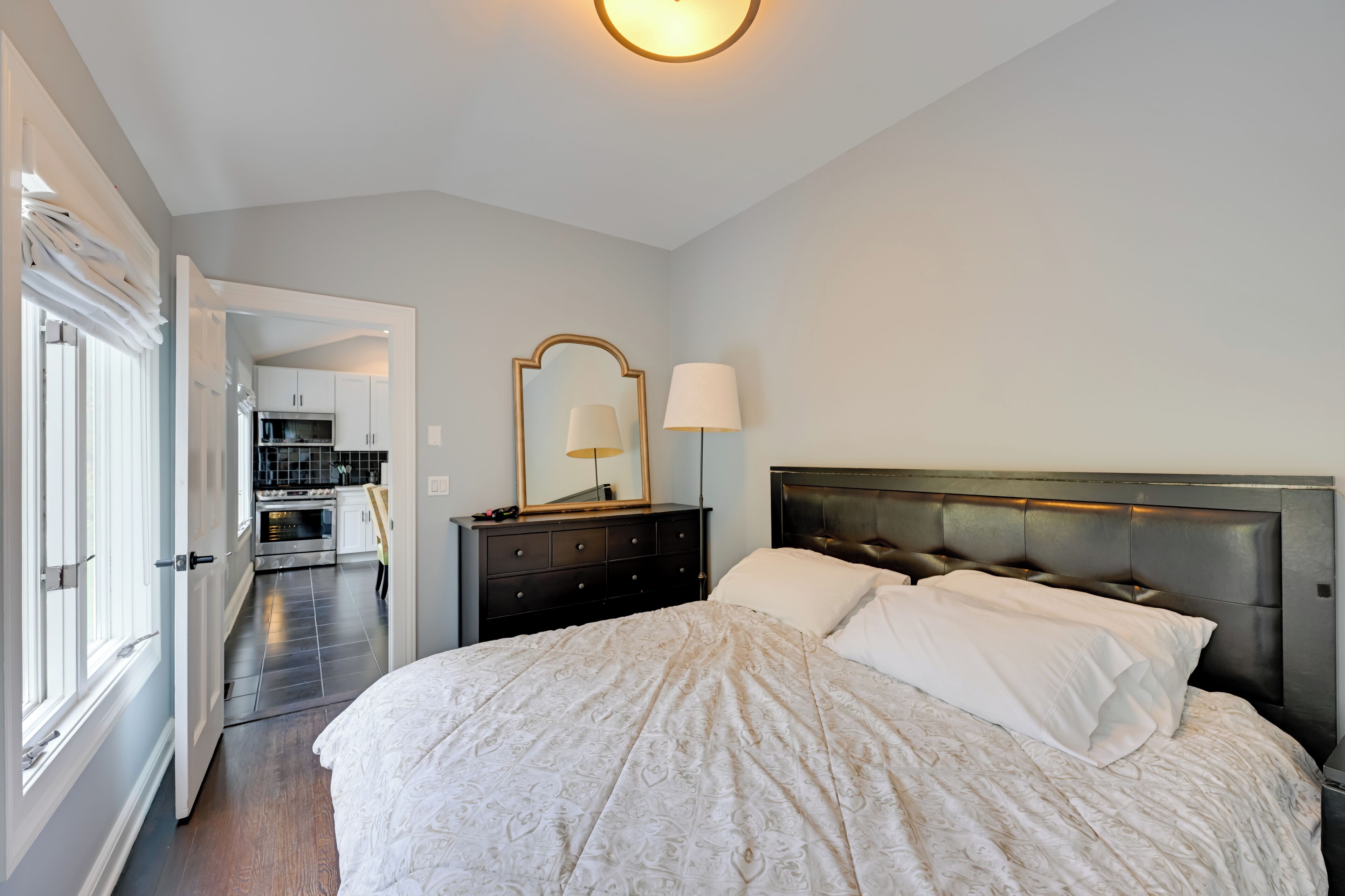 Une chambre avec un grand lit, une table de chevet avec une lampe, une commode et un miroir. On peut apercevoir une cuisine à travers une porte ouverte.