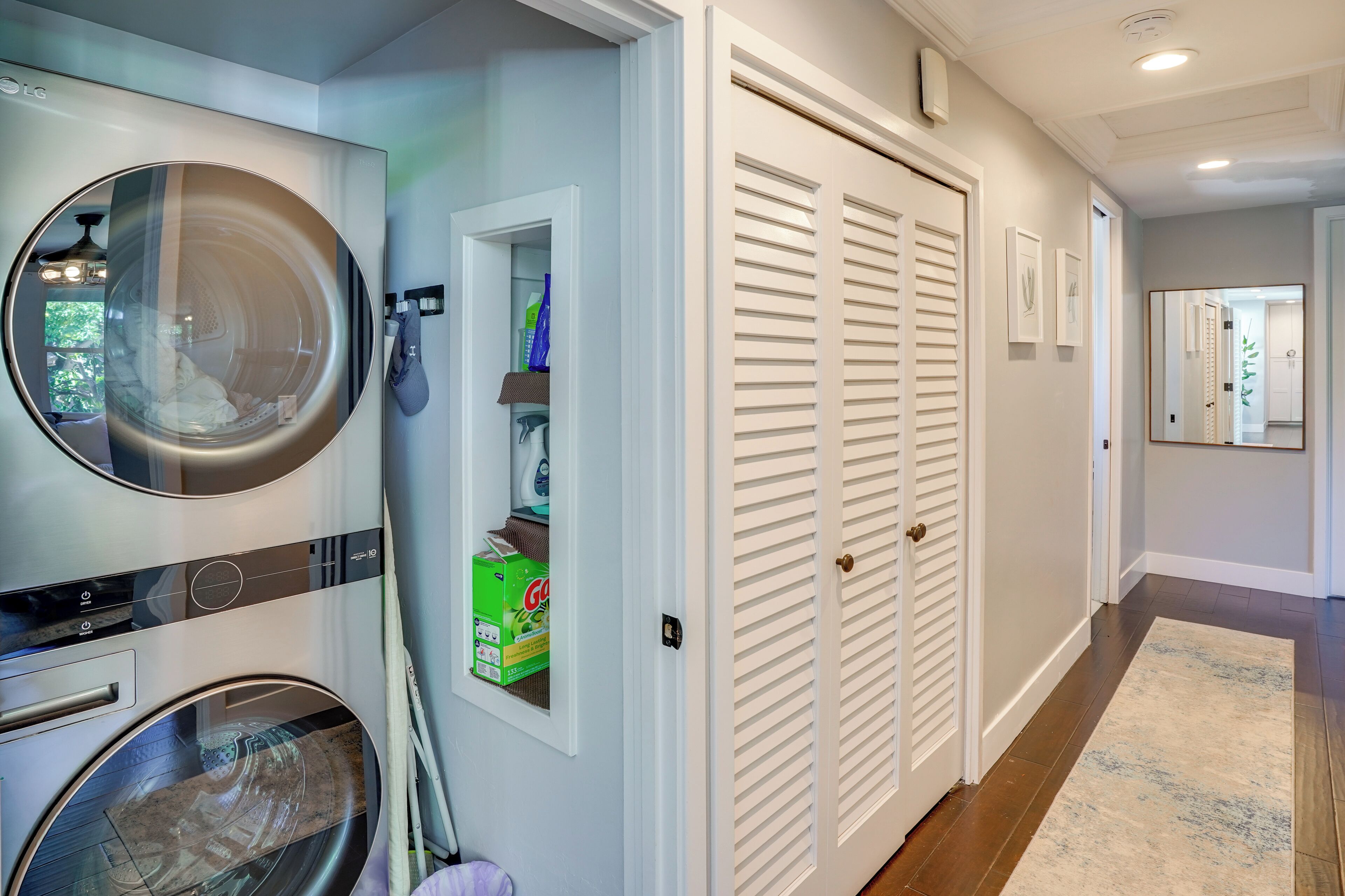 A laundry room with a front-loading washer and dryer, a storage closet, and a hallway with a wooden floor and a rug.