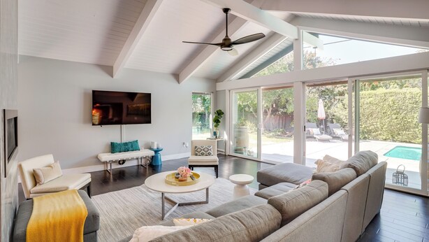 A modern living room with a large sofa, a coffee table, and a TV mounted on the wall. There is a sliding glass door leading to an outdoor area with a pool.