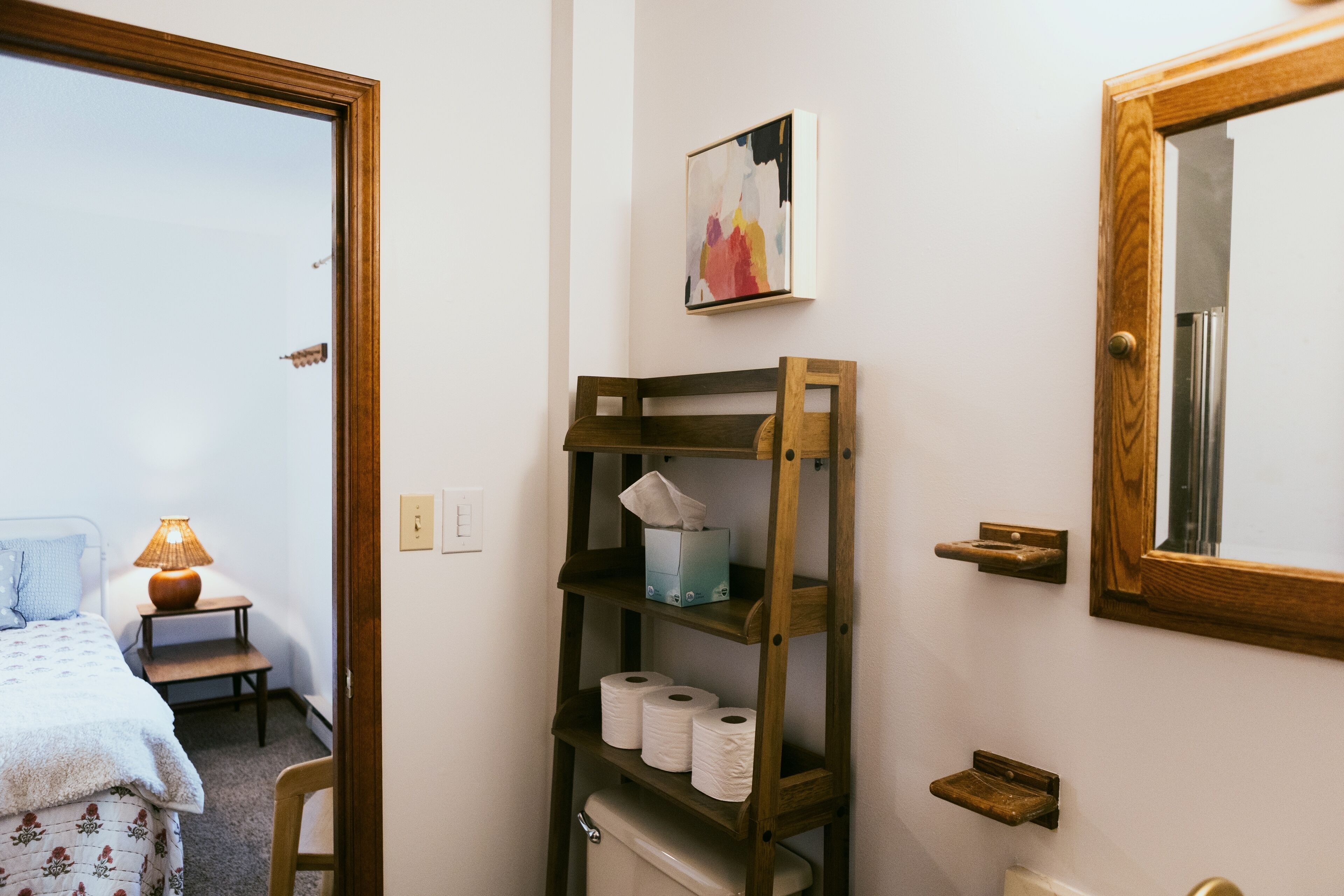 A bathroom with a wooden shelf holding toilet paper and a tissue box, a mirror, and a painting on the wall.