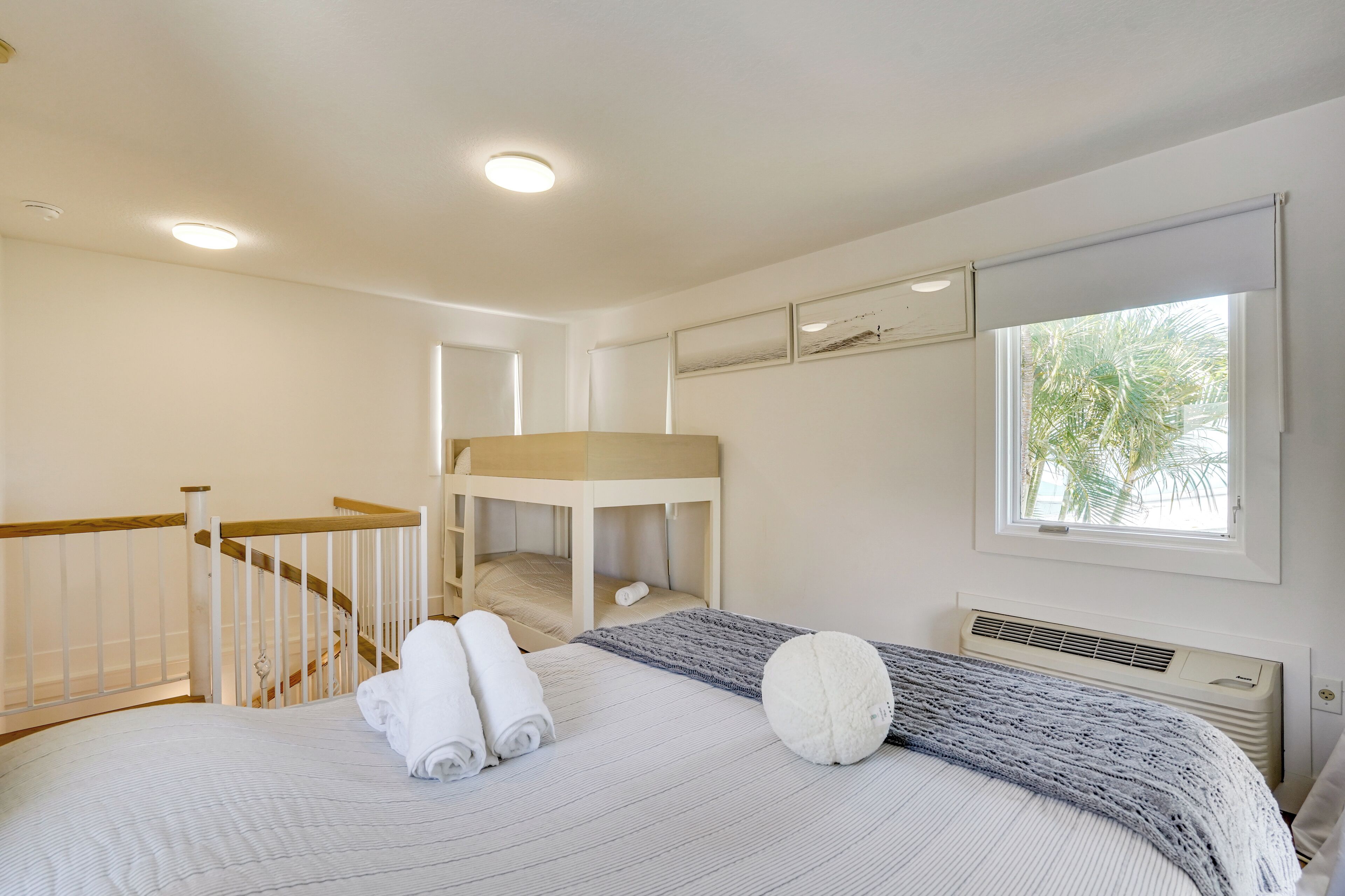 A bedroom with a bunk bed, a bedside table, a window with a view of palm trees, and a ceiling fan.