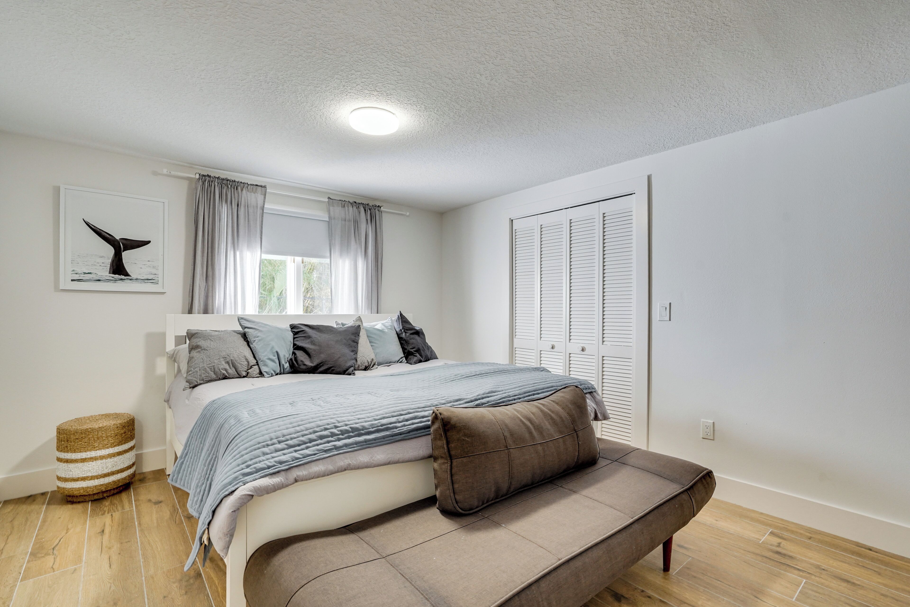 A bedroom with a bed, a bench, a window with curtains, a wall-mounted artwork, and a door with shutters.