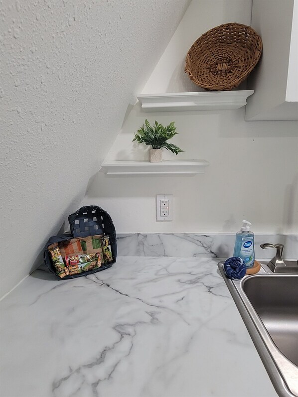 Una cocina con encimeras de mármol blanco, una bolsa negra de bocadillos, una pequeña planta en maceta, una cesta tejida y una botella de jabón para manos Dial.