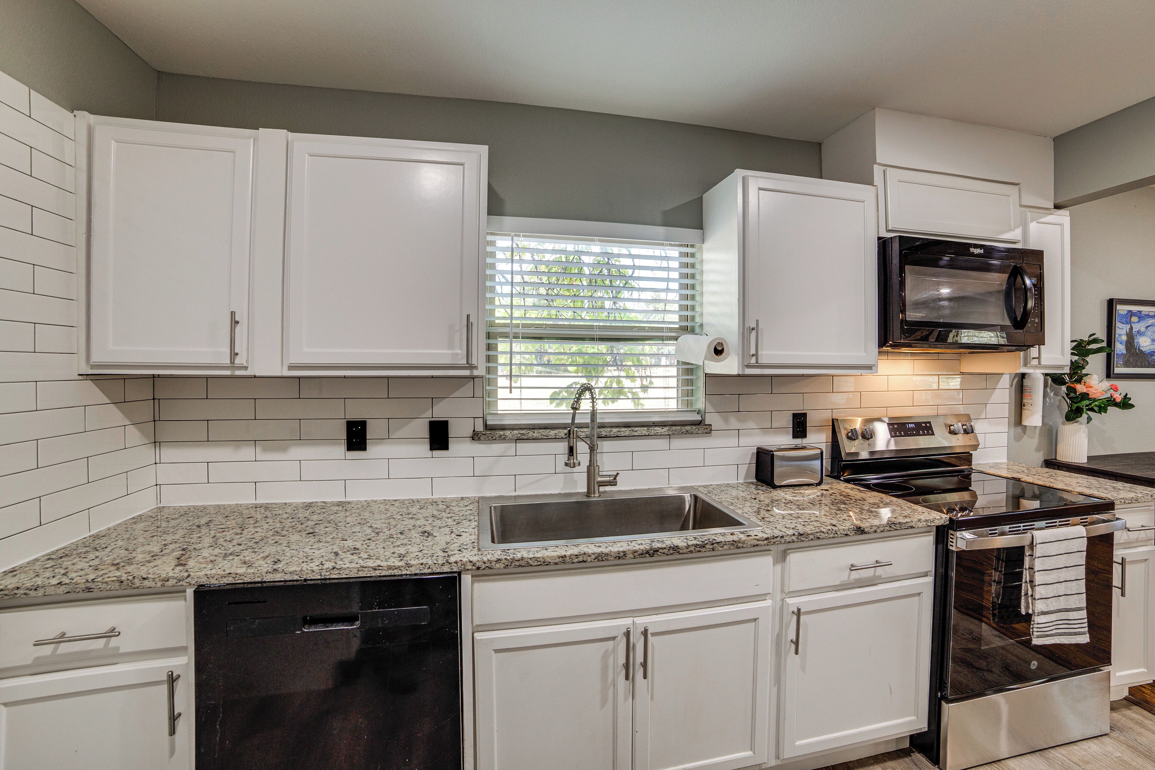 Una cocina moderna con armarios blancos, encimera de granito, electrodomésticos de acero inoxidable y una ventana con persianas.