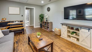 A living room with a sofa, coffee table, wooden flooring, a TV, a bookshelf, and a door leading to another room. - Walk to East Texas A&m! Home w/ Pet-friendly Yard (Commerce)