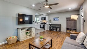A modern living room with a flat-screen TV, a grey sofa, a wooden coffee table, and a kitchen in the background. - Walk to East Texas A&m! Home w/ Pet-friendly Yard (Commerce)