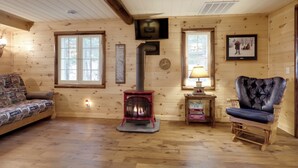 Ein gemütliches Blockhaus-Interieur mit Holzboden, einem roten Holzofen, einer Couch, einem Ledersessel und einem kleinen Tisch mit einer Lampe.