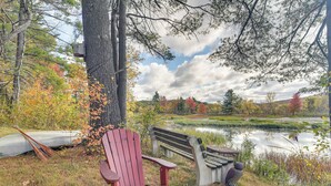 Un paisaje tranquilo junto al lago, con una silla Adirondack roja y un banco de madera cerca de un estanque, rodeado de árboles y follaje otoñal.