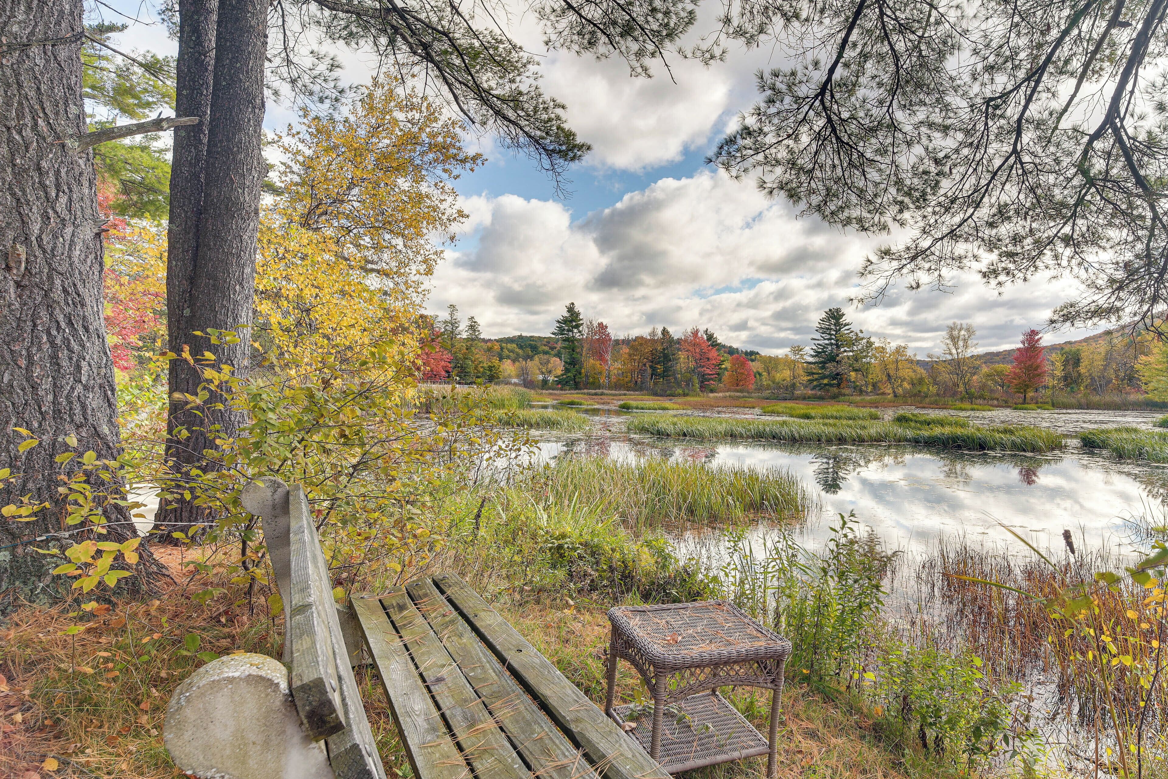 A wooden bench and a small table by a calm pond surrounded by trees with autumn foliage.