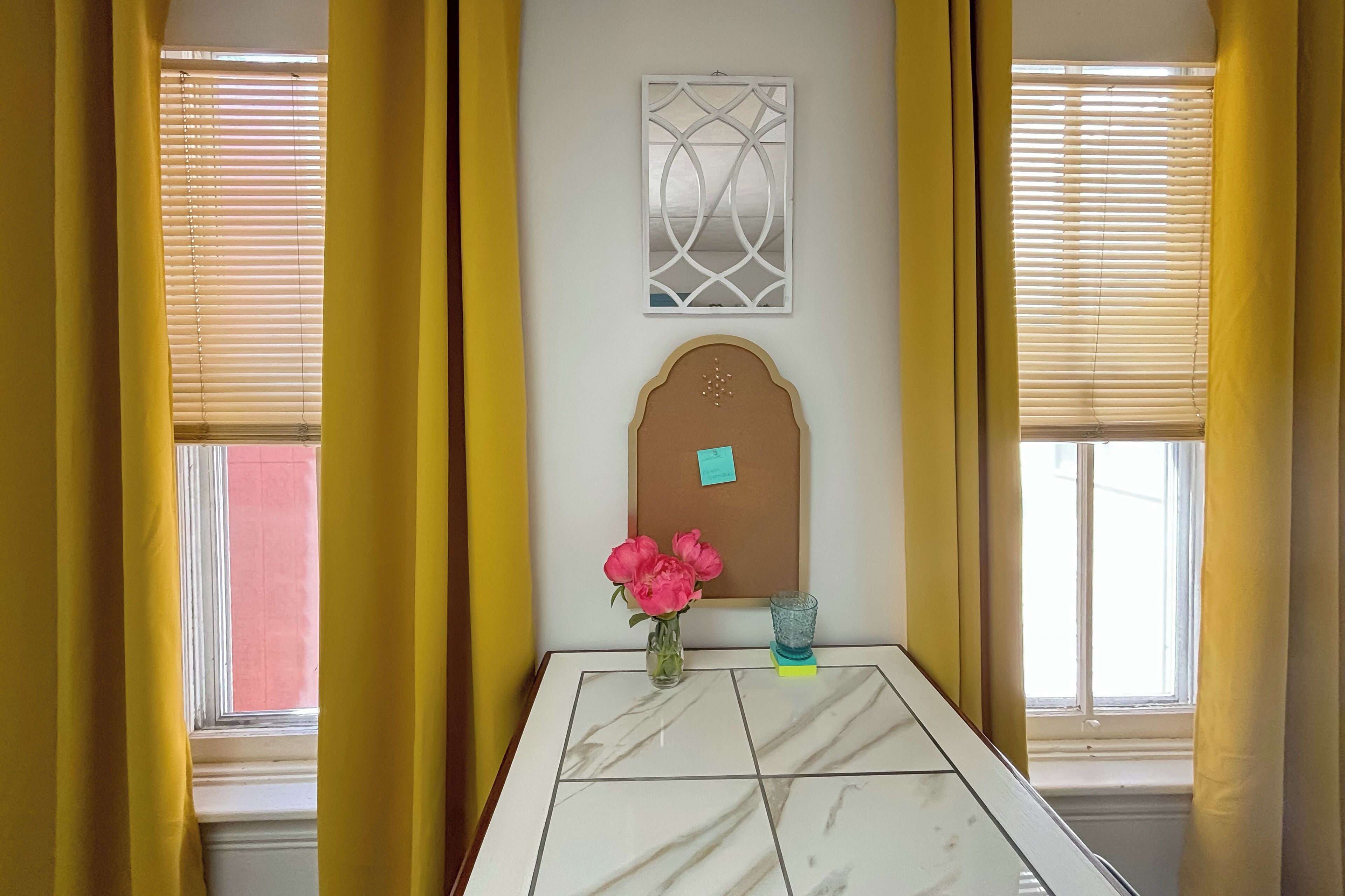 A room with a marble-tiled table, a vase with pink flowers, a blue glass, and a bulletin board with a note.