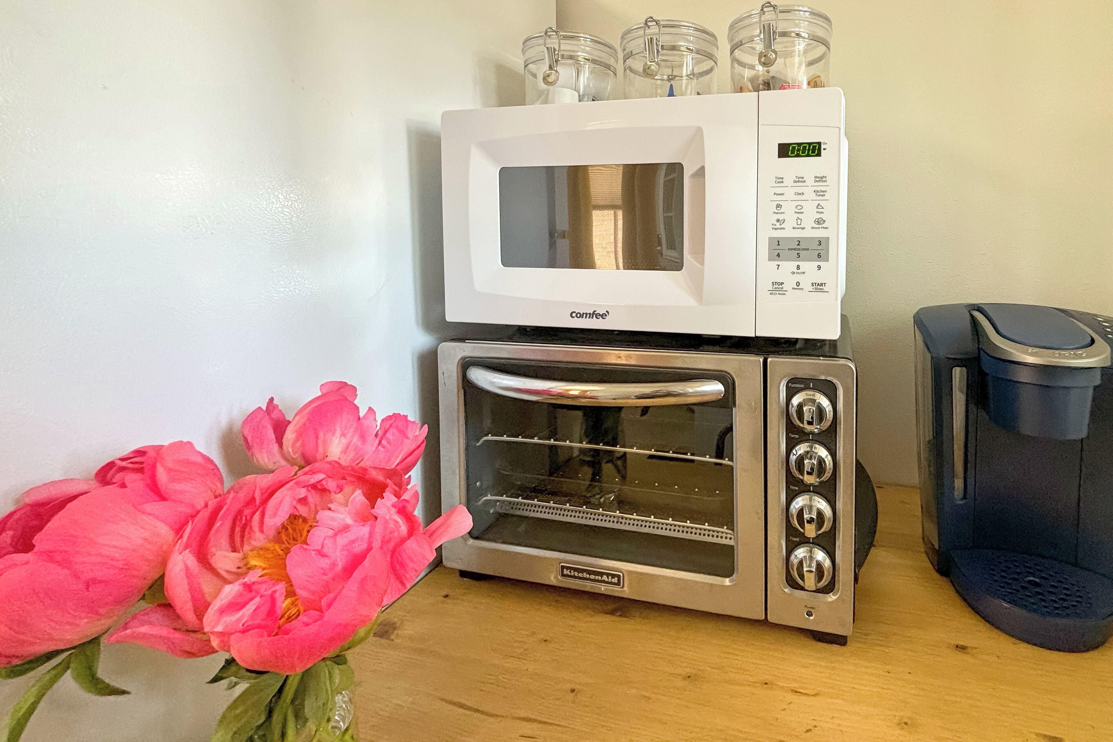 A kitchen with a microwave and a toaster oven, a trash bin, and a vase with pink flowers.