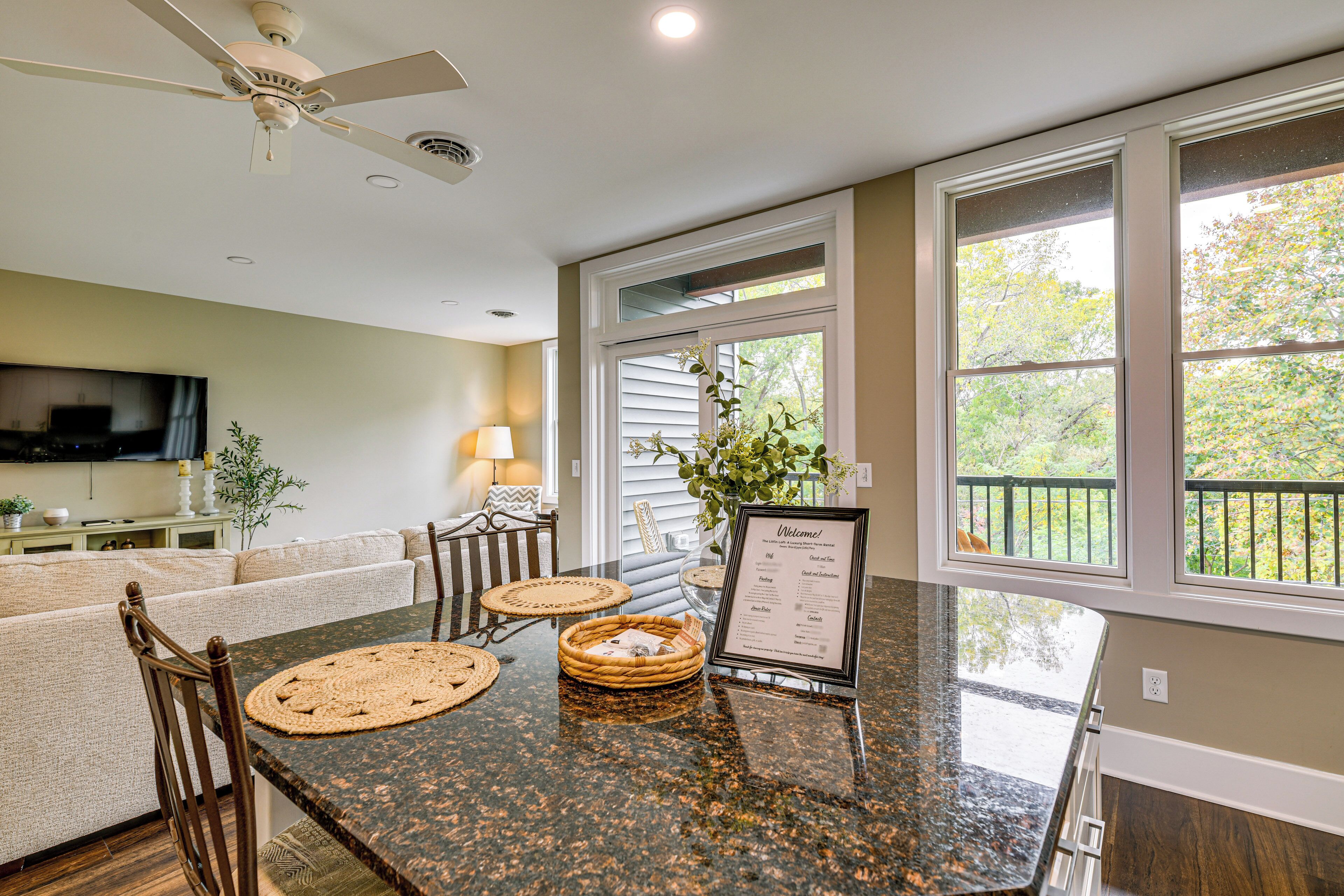 A modern kitchen with a granite countertop, a dining table with chairs, a ceiling fan, and a view of the outdoors through large windows.