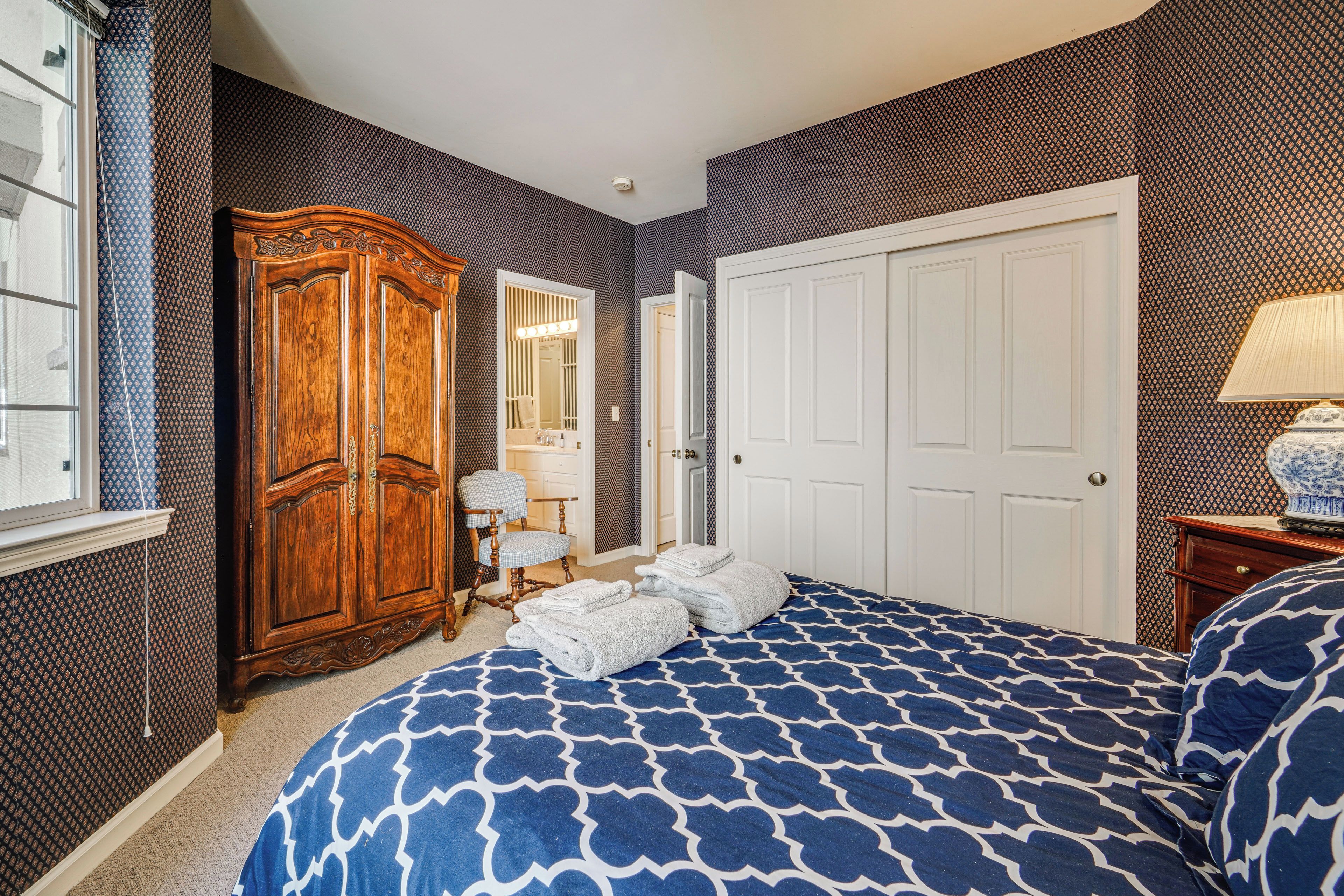 A bedroom with a patterned bedspread, a wooden wardrobe, a chair, and a mirror.