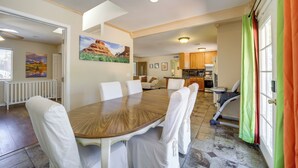 Un coin repas avec une table en bois et des chaises blanches, une cuisine en arrière-plan et une vue sur l’extérieur par une porte.