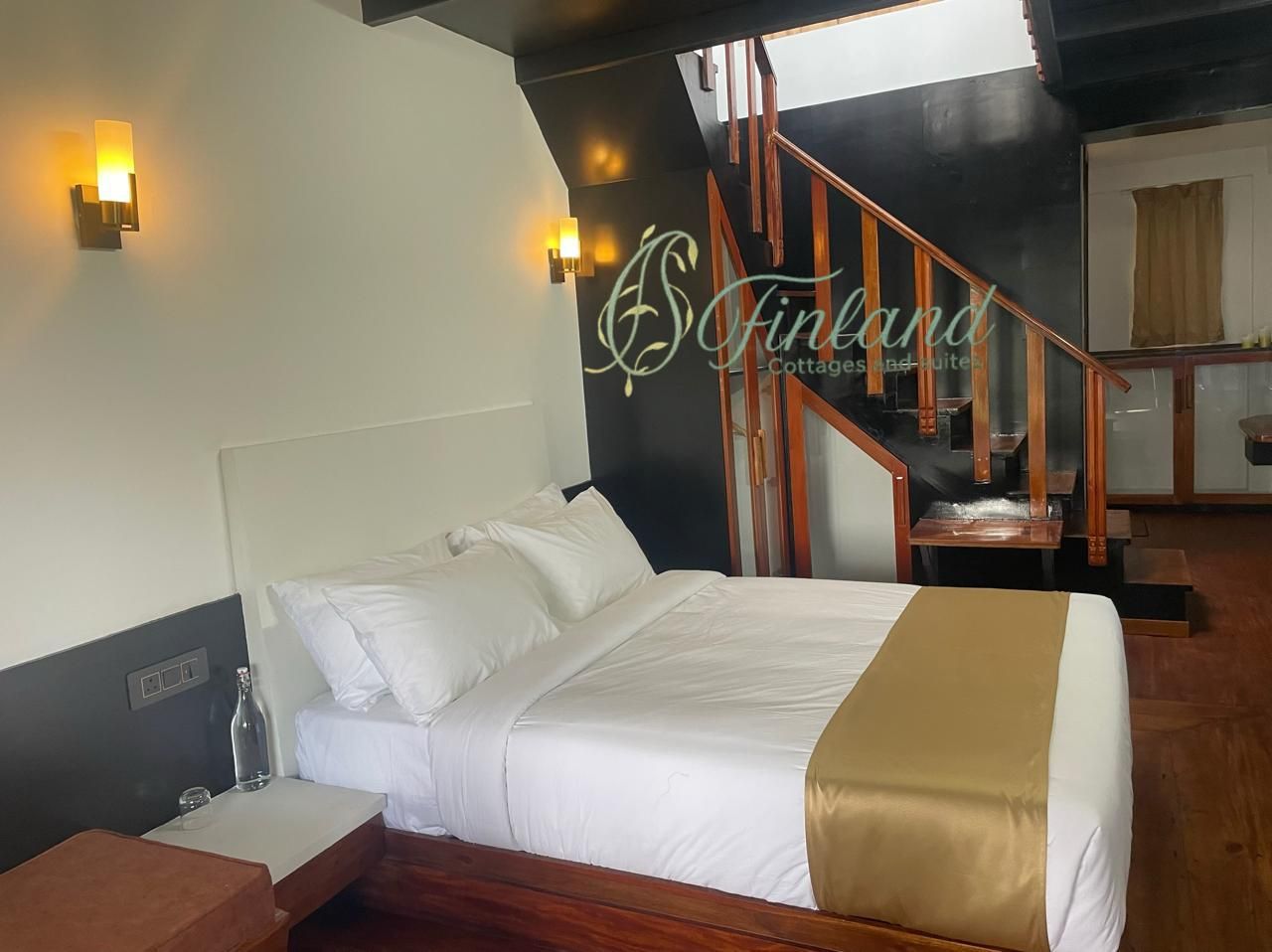 A hotel room with a bed, a nightstand, a bottle, and a glass. There is a staircase with wooden railings and a wall with a logo.