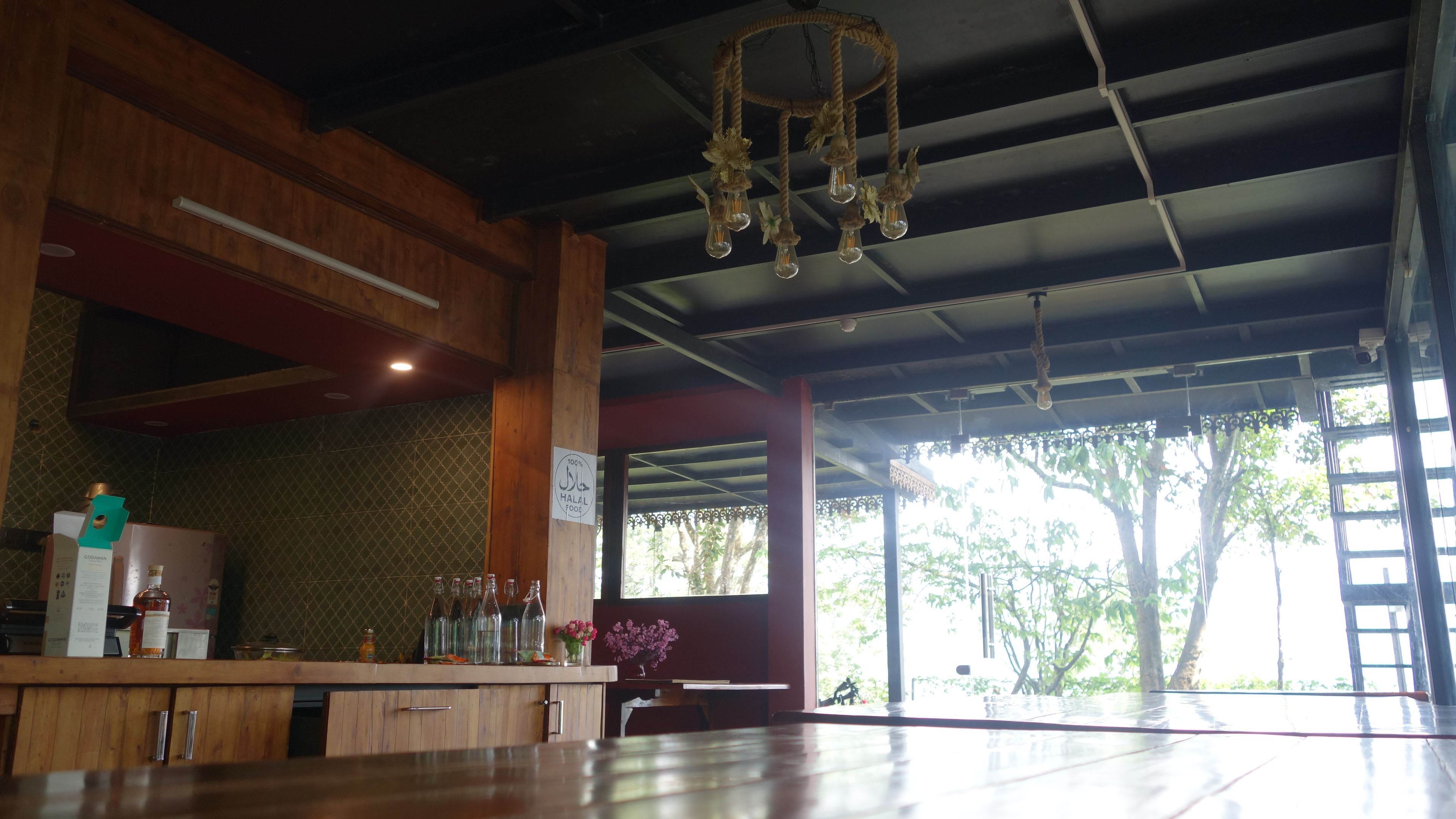 A restaurant interior with a wooden counter, hanging light fixtures, and a view of trees through large windows.