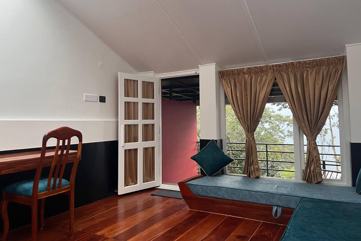 Quarto com piso de madeira, escrivaninha com cadeira, janela com cortinas e vista para áreas verdes.