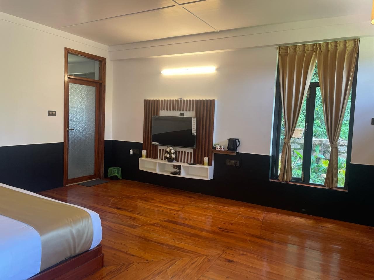 Une chambre d’hôtel avec un lit, un téléviseur fixé sur un panneau en bois, une fenêtre avec des rideaux et une petite étagère avec quelques objets.