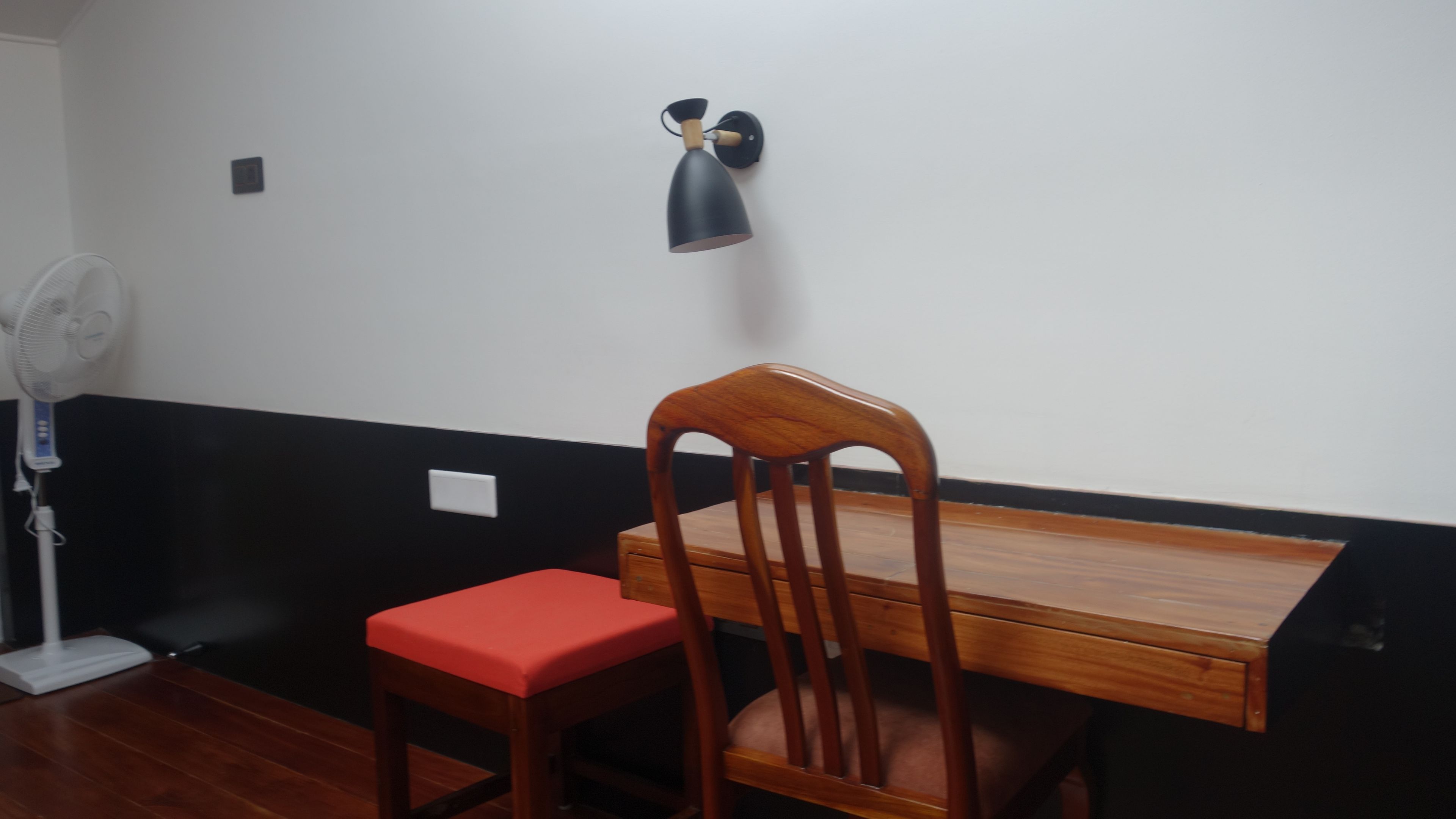 A room with a wooden desk, a red cushioned chair, a wall-mounted lamp, and a standing fan.