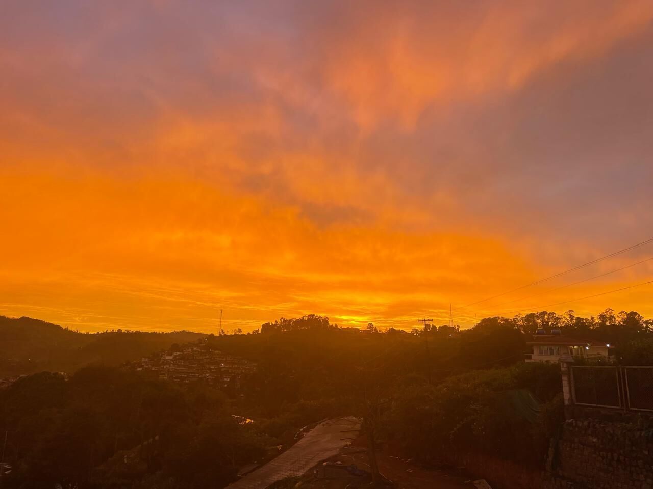 Un coucher de soleil sur un paysage vallonné, avec une route et des bâtiments visibles au loin.