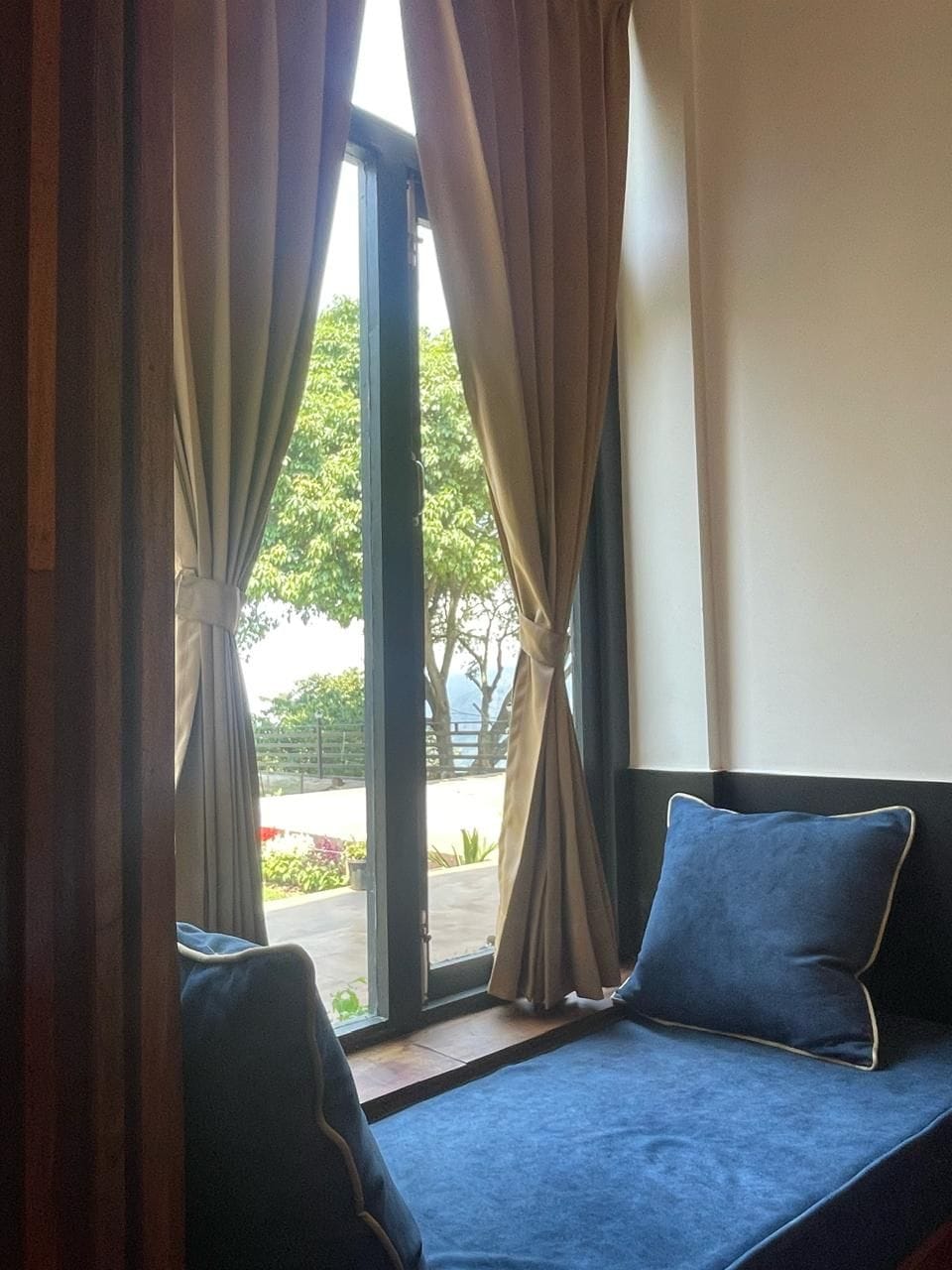 A room with a window, curtains, a sofa with a cushion, and a view of greenery outside.