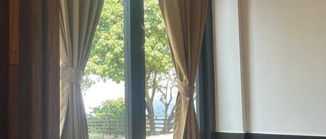 A room with a window, curtains, a sofa with a cushion, and a view of greenery outside.