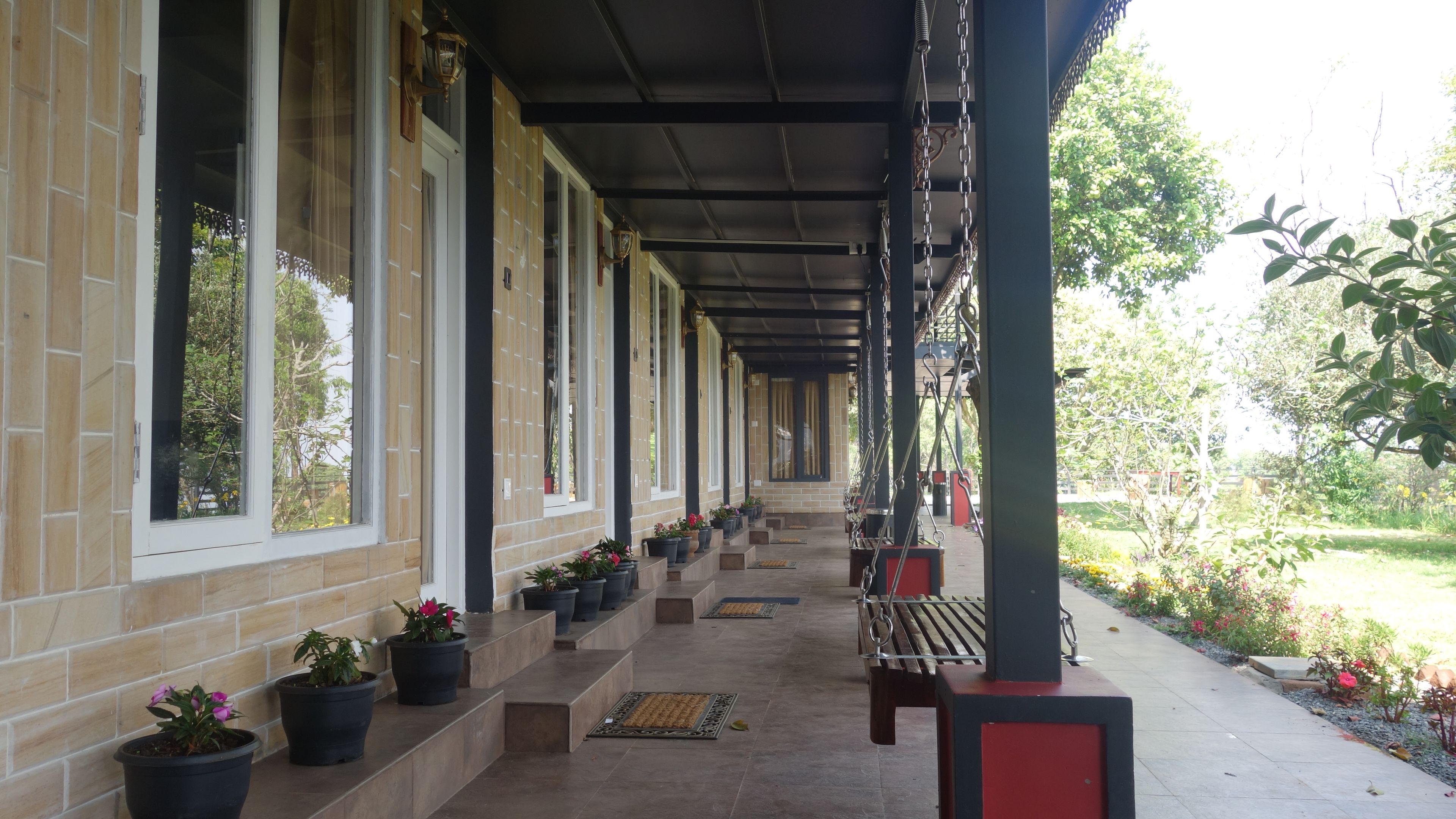 A covered outdoor area with a row of potted plants, a bench, and a pathway leading to a building entrance.
