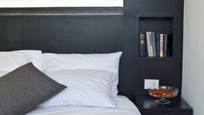 A neatly made bed with white and gray pillows, a small black nightstand with a bowl of snacks, and a bookshelf with books. - Luxury attic with jacuzzi in Navigli (Milano)