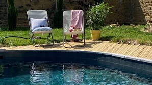 Una piscina transparente con dos sillas reclinables blancas y una planta amarilla en maceta sobre un muelle de madera.
