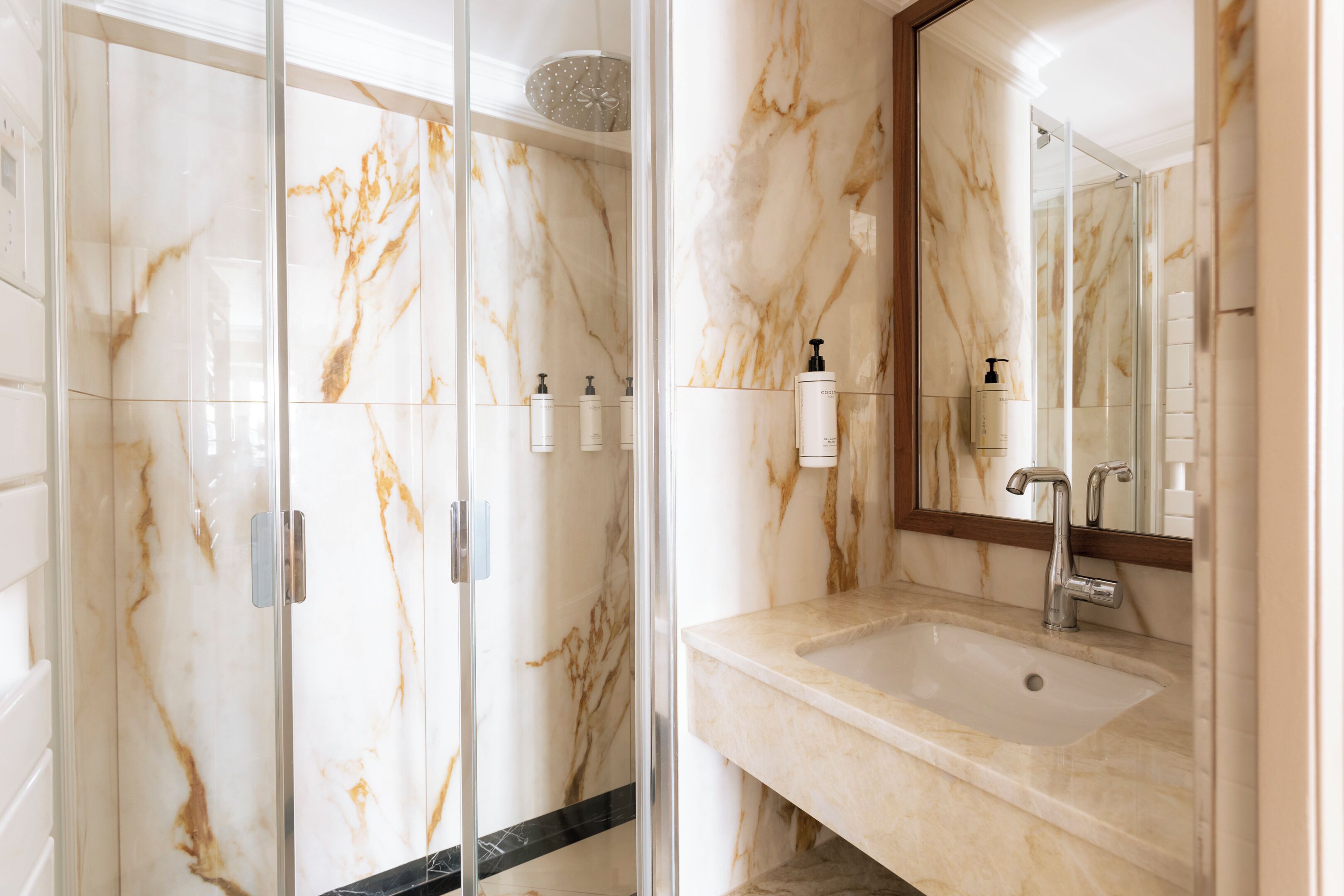 Une salle de bain avec une douche à parois de verre, un mur recouvert de carreaux de marbre, un lavabo avec un miroir et des articles de toilette.