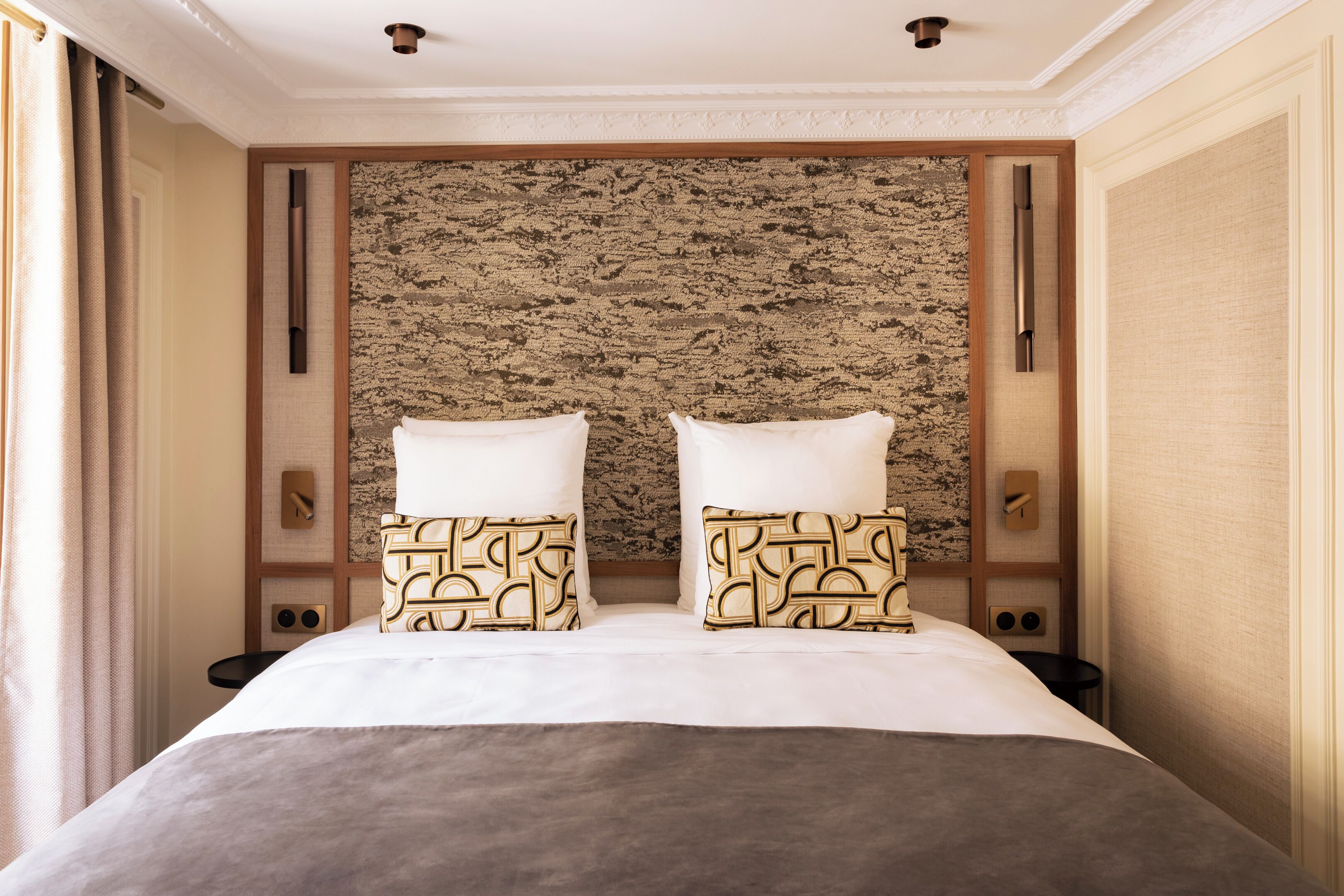 A hotel room with a large bed, two pillows, a textured headboard, and a window with curtains.