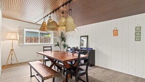 A dining area with a wooden table, dark chairs, and a bench. There are hanging lights, a floor lamp, and a window with blinds. - Indio Poolside Retreat Getaway by Navigate Stays (Indio)