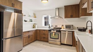A modern kitchen with stainless steel appliances, wooden cabinets, a built-in oven, and a window providing natural light. - Indio Poolside Retreat Getaway by Navigate Stays (Indio)