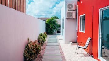 Un étroit couloir extérieur, avec un bâtiment rouge sur la droite, un bâtiment blanc sur la gauche, et un sentier bordé de plantes.