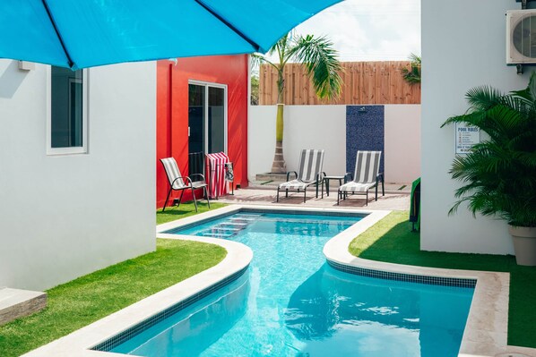 A swimming pool with a tiled edge, surrounded by a small grassy area and a red building.
