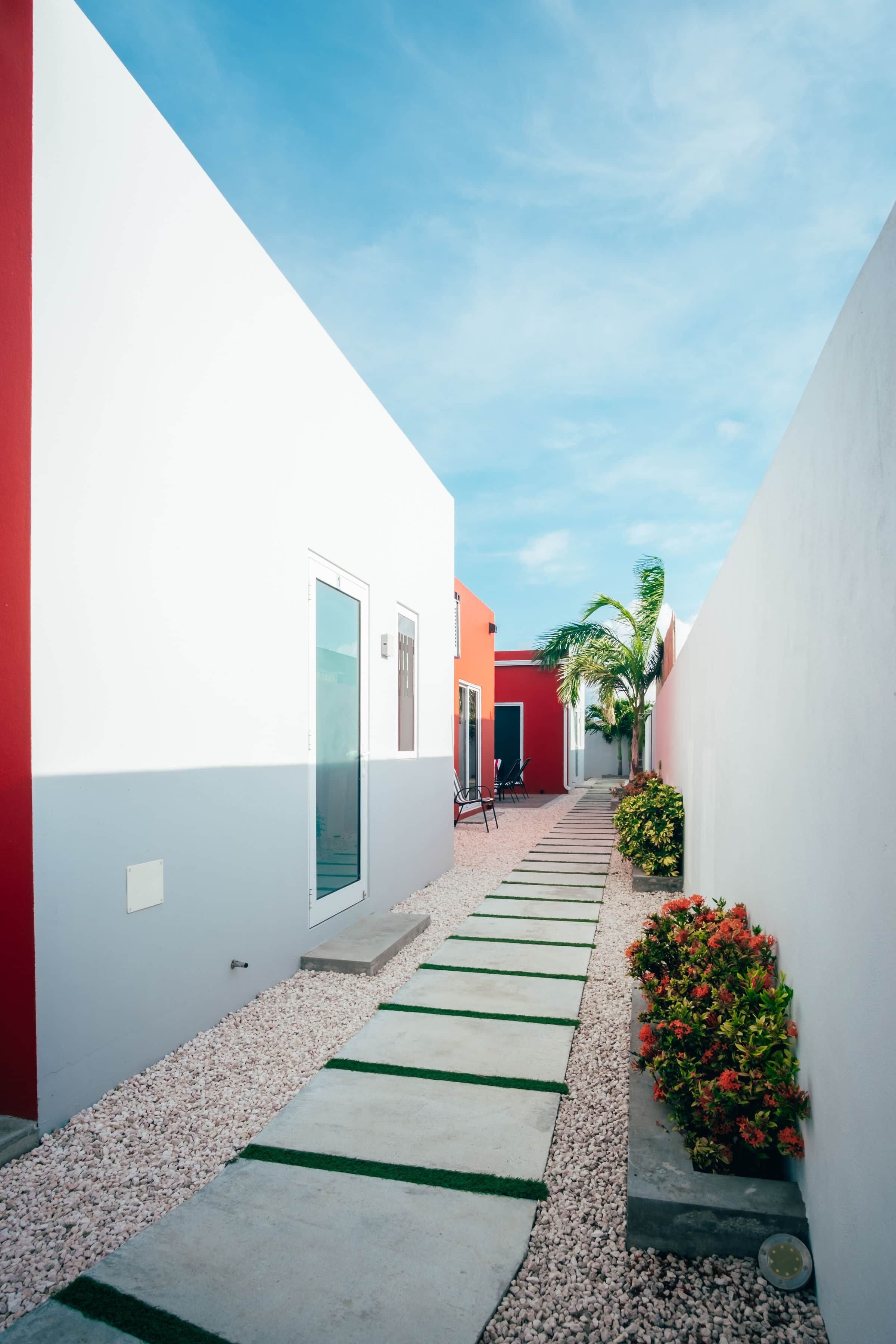 Ein schmaler Weg führt zu einem Gebäude mit roter Fassade, umgeben von weißen Mauern und blauem Himmel.