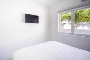 Ein Zimmer mit einem Bett, einem an der Wand montierten Fernseher und einem Fenster mit Blick auf Grünflächen.