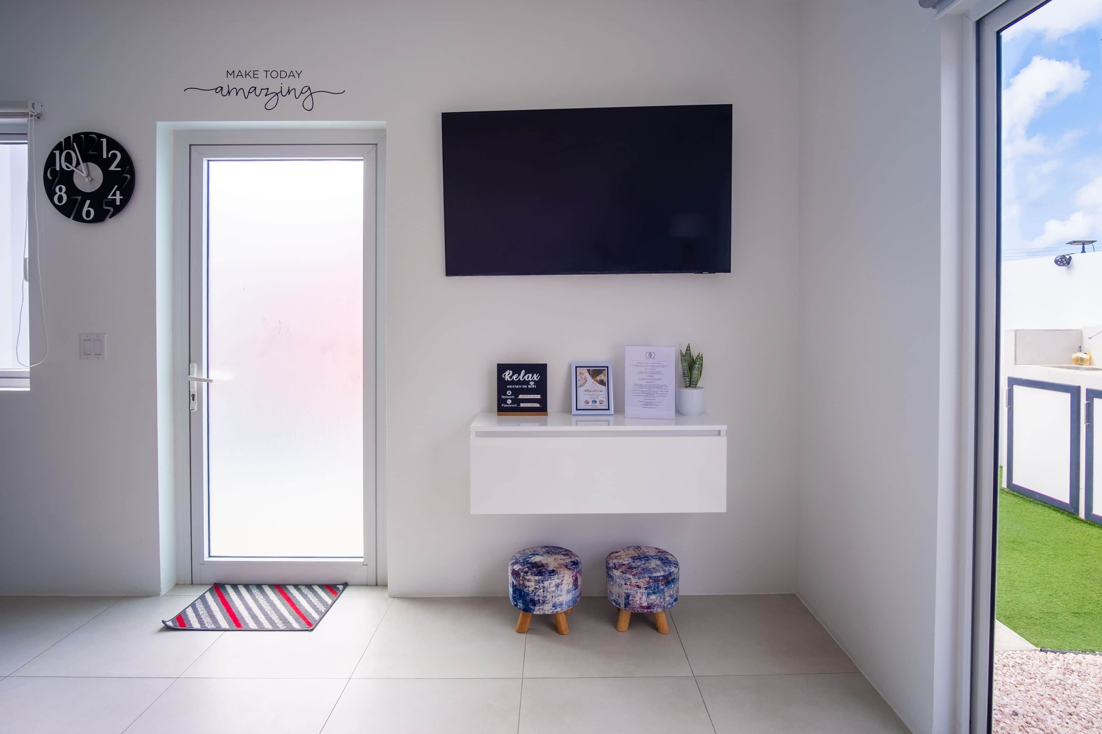 Ein modernes Wohnzimmer mit einem flachbildfernseher an der Wand, einer weißen Tür mit einer Uhr und einem kleinen Regal mit Dekorationselementen.
