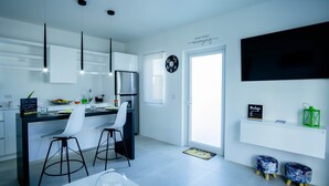 Une cuisine moderne avec des meubles blancs, un four encastré et une grande fenêtre. On y trouve deux tabourets, une table blanche et un saladier en verre sur le plan de travail.