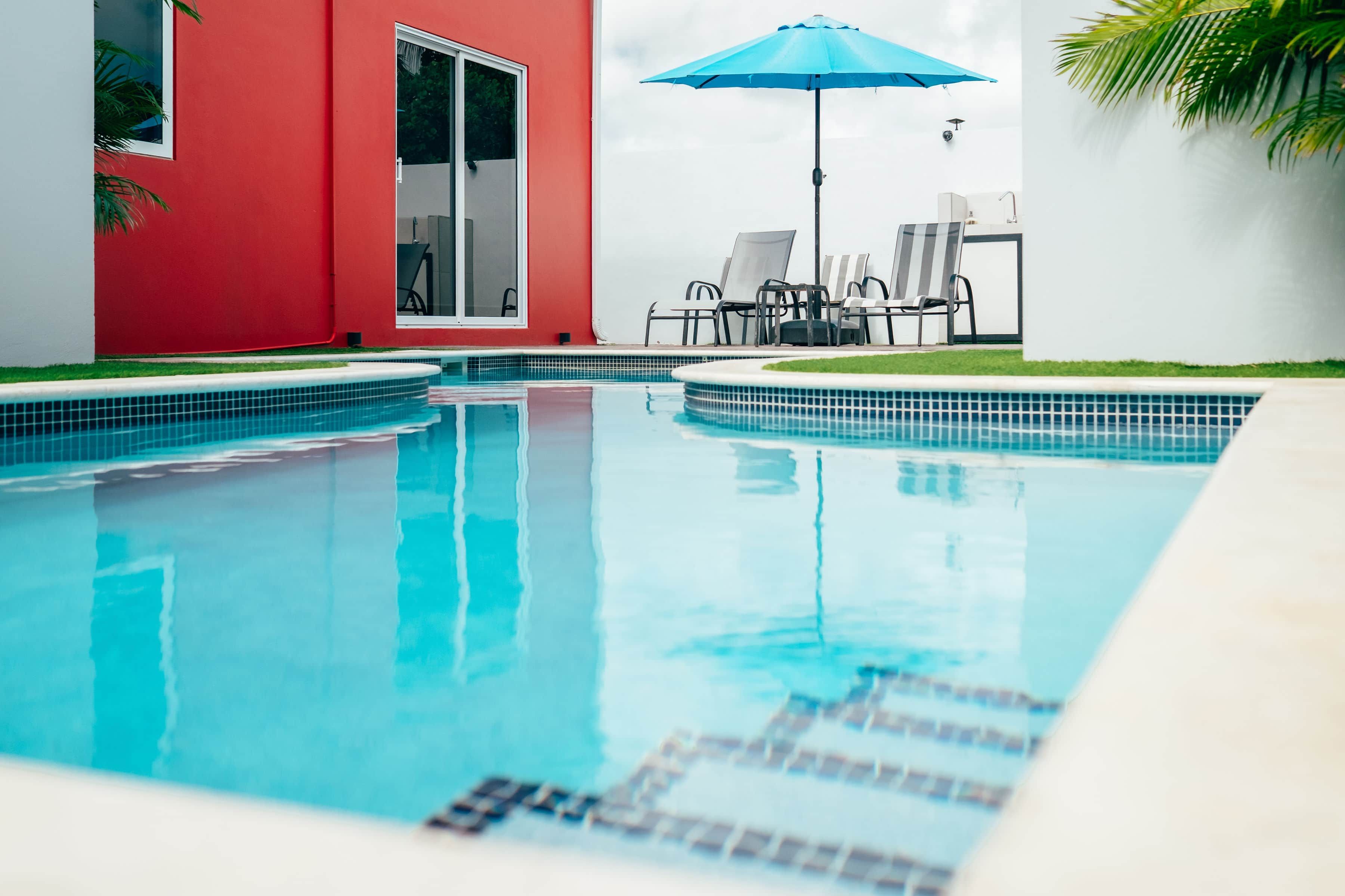 A swimming pool with a tiled edge, a red building, and a blue umbrella.