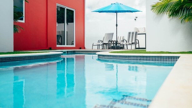 Ein Schwimmbecken mit einer gefliesten Kante, ein rotes Gebäude und ein blauer Sonnenschirm.