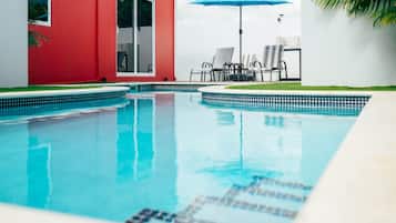 Une piscine avec un bord carrelé, un bâtiment rouge et un parasol bleu.