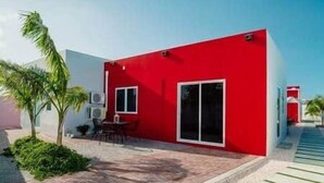 Un bâtiment moderne de couleur rouge, doté d’une grande porte-fenêtre coulissante, d’un petit espace patio avec une table et des chaises, et d’un palmier.