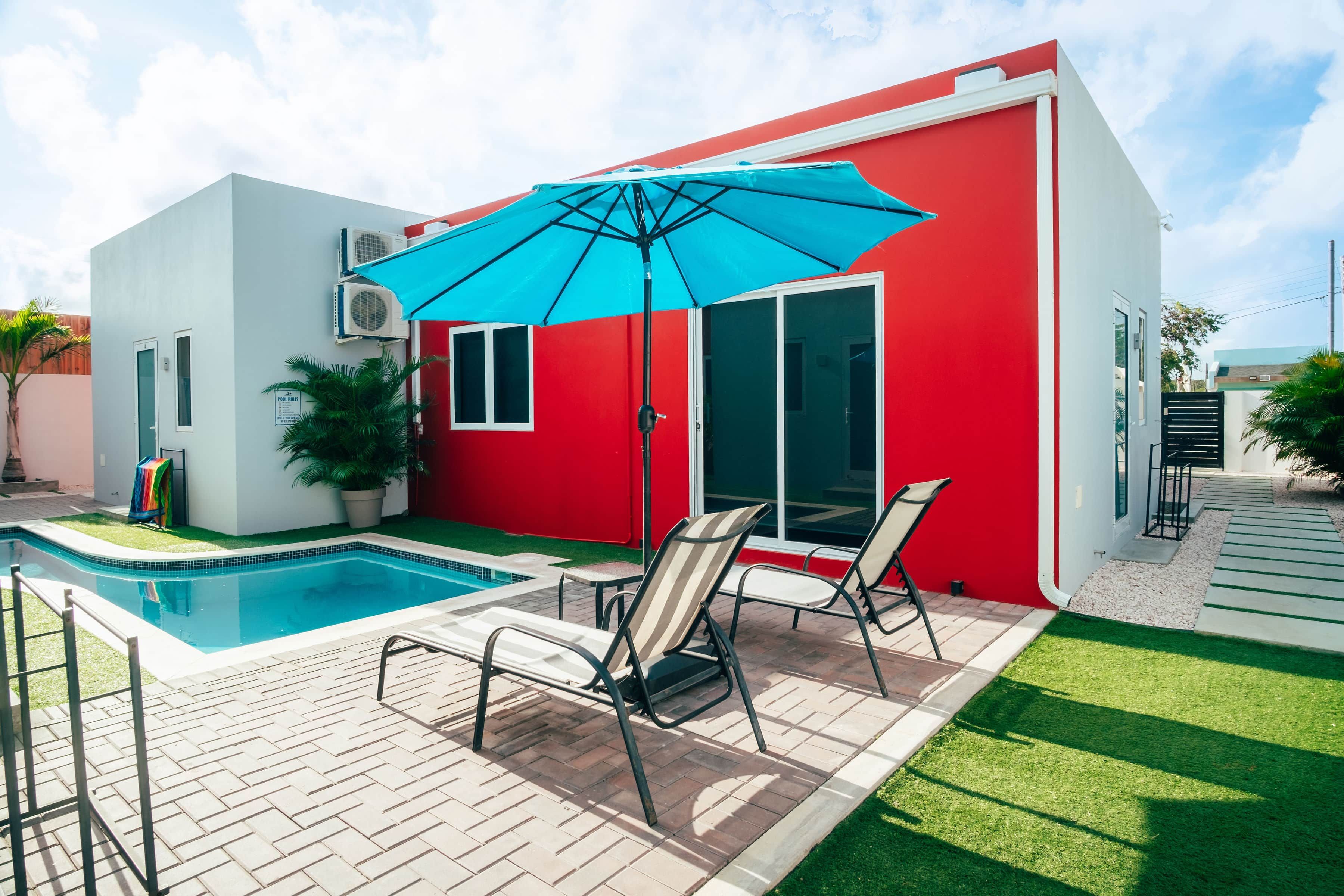 Ein modernes Haus mit rotem Anstrich, einem Schwimmbad und einer Terrasse mit Stühlen und Sonnenschirm.