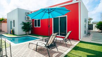 Ein modernes Haus mit rotem Anstrich, einem Schwimmbad und einer Terrasse mit Stühlen und Sonnenschirm.