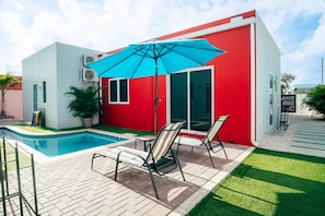 Une maison moderne à l’extérieur rouge, dotée d’une piscine et d’un patio aménagé avec des chaises et un parasol.