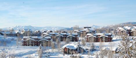 Un centro turístico cubierto de nieve con varias edificaciones de estilo chalet, una pista de esquí al fondo y un cielo despejado.
